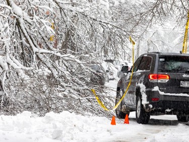 A vehicle on a snow-covered street right beside fallen branches with yellow caution tape and orange cones visible