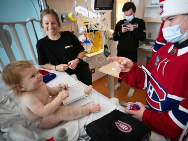 A baby in a hospital bed smiles while Cole Caufield points back smiling behind a mask