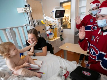 A baby on a hospital bed waves to two Canadiens players