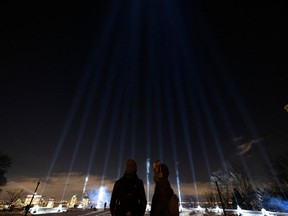 Two people stand in the dark in front of 14 beams of light shining into the air at a lookout with the city skyline in the background