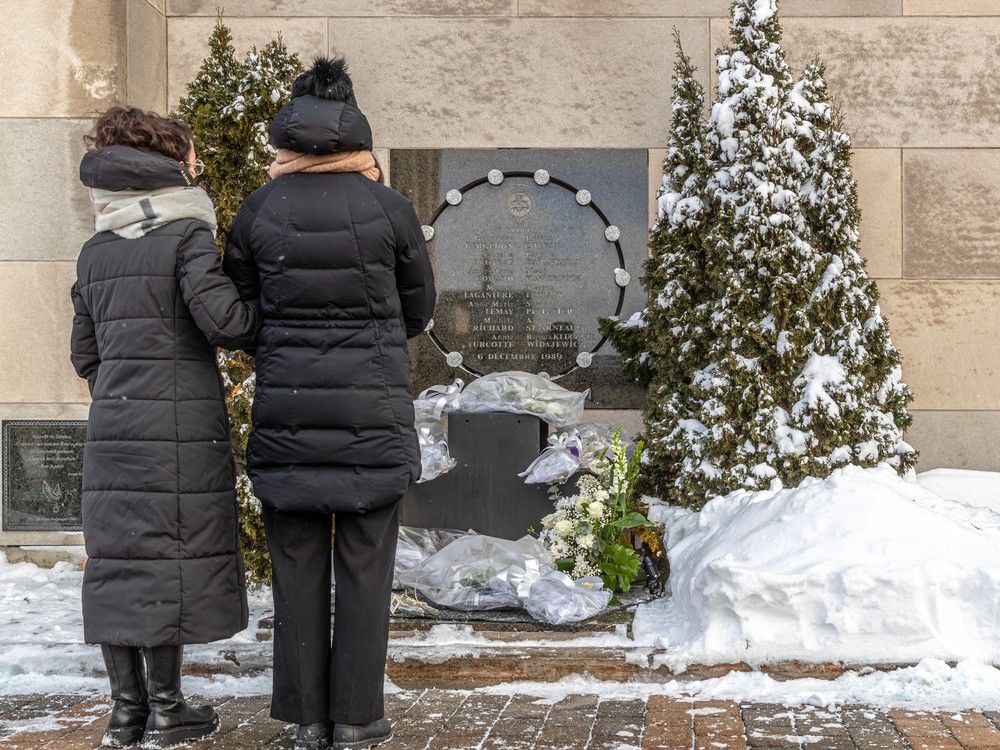 two women, seen from behind, stand before a plaque where people have placed wreaths of white roses.