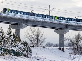 A light rail train is seen on an elevated track above ground covered in snow with a few trees and shrubs.