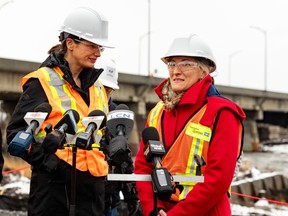 Two women in hard hats stand outside in front of media microphones with a bridge in the background