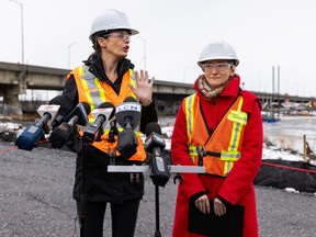 Two women in hard hats and reflective vests stand in front of microphones with a bridge visible in the background