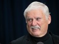 A man with white hair and a moustache is smiling against a black backdrop.