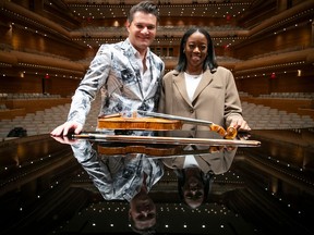 A man and woman pose behind a violin on top of a piano in a concert hall.