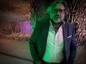 A man poses under purple and green lights. He is wearing a suit and glasses and has long grey hair.