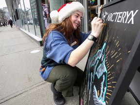 A woman in a Santa hat kneels down to write on a sign outside a store.