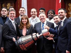 Politicians smile holding up the Grey Cup with football players behind them in the Red Room