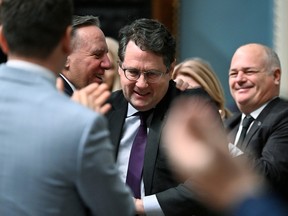 Bernard Drainville shakes hands with François Legault while smiling in the Blue Room at the National Assembly while caucus colleagues applaud around them
