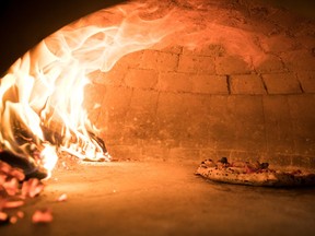 Flames emanate from wood logs on one side of an arched stone oven while a pizza cooks on the other side
