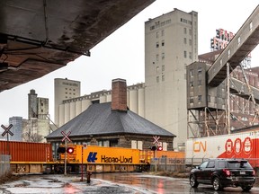 A freight train passes buildings on its way under an overpass.