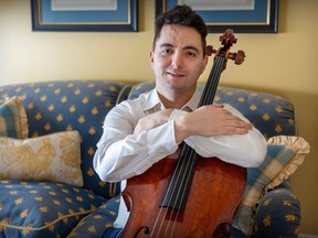 A man smiles while holding a cello as he sits on a couch.