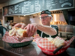 A man prepares a submarine sandwich in a restaurant kitchen. He is putting the sandwich into a basket.
