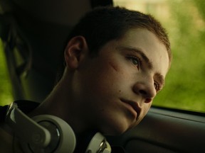 A sad teenage boy looks out a window. He has headphones around his neck.