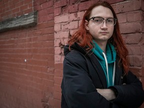 A person with long, red hair poses in front of a brick wall, wearing a black jacket with a turquoise hoodie underneath.