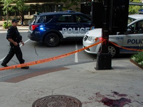 A cop walks near two police vehicles and orange tape next to a sidewalk that has a bloodstain on it.