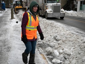 A woman in an orange safety vest walks on a sidewalk in front of snow-clearing trucks