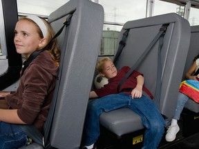Two children on a school bus: One is looking toward the driver, the other is stretched out on the bench, sleeping with his head on a soccer ball.