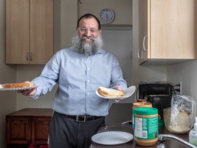 Rabbi Benyamin Bresinger smiles as he holds paper plates with sandwiches in each hand in a kitchen, with jars of peanut butter on the counter.