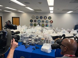 Cameras are seen taking picture of tables with seized drugs, money and weapons on them.