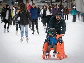 A man pushes a child in a rolling chair on a skating rink in the foreground of a group of skaters.