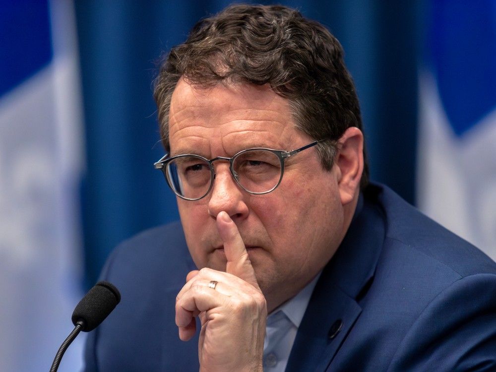 quebec education minister bernard drainville is seen in this closeup photo of him beside a microphone, holding his index finger to his mouth while squinting.