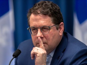 Quebec education minister bernard drainville is seen in this closeup photo of him beside a microphone, holding his index finger to his mouth while squinting.
