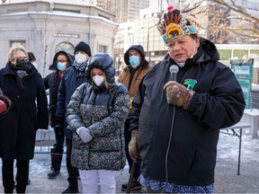 a man wearing a traditional Indigenous headdress is holding a microphone and speaking at a memorial, flanked by other people attending.