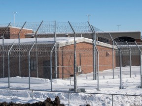 A prison is seen from the outside, with a tall fence with barbed wire in front of a pale brown brick building. There is snow on the ground.