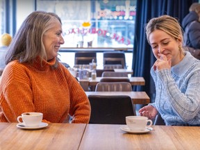 A woman in an orange sweater with grey hair is sitting in a cafe, with a window in the background, beside a woman in a grey sweater and blonde hair. Both have coffee cups sitting on a table in front of them.
