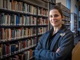 A woman with brown hair tied back, wearing a black blazer-type coat stands cross-armed in front of a bookshelf filled with multi-coloured books.