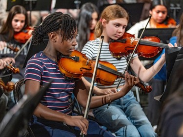 Two students practice violin with their peers in the background.