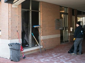 A worker looks at a broken window of a reception hall. A broom and trashcan are against the wall of the building.