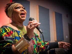 A woman in a colourful dress and headband is holding up a microphone and singing into it in front of a wall painted with vertical rectangles.