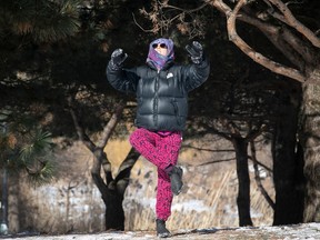 A woman in bright pink pants has her arms raised and one leg lifted as she exercises in a park.