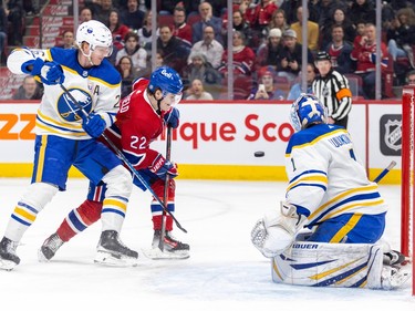 The puck is in the air between Canadiens' Cole Caufield, whose stick is being held back by a Sabres player's, and the Sabres goaltender