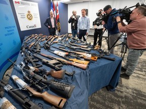 A table is covered in firearms. Photographers and cameramen are surrounding the table.