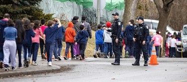 Students walk down the sidewalk in a row as two police officers observe them.