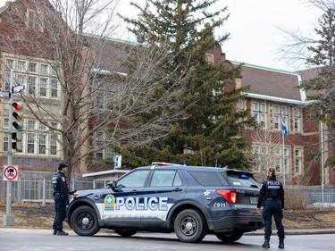 Two police officers flank a police cruiser in front of a large brick building, blocking off the street..