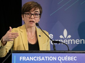A photo of a woman with short brown hear wearing glasses and a yellow blazer, standing at a podium and gesturing with her right hand. The podium says 'francisation quebec' and there's a blue backdrop behind her.
