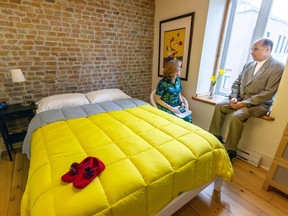 A man sits on a windowsill talking with a woman in a chair in bedroom with a brick wall and bright yellow comforter on a bed.