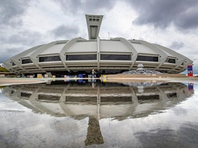 The Olympic Stadium in Montreal is seen from eye level on a cloudy/rainy day. It is being reflected in the water on the ground in front of it, and the sky behind it is cloudy.