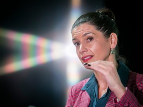 A woman wearing a pink blazer is staring into the camera gesturing with her hand in front of her chin. The background is mostly black, with a rainbow-coloured reflection forming the shape of a star behind her.