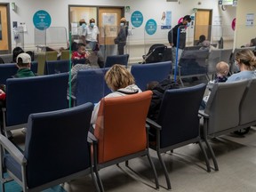 People sit in a hospital waiting room. The chairs have plastic dividers between them.