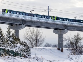A white, green and black train is seen travelling left on a concrete overpass. The ground beneath is snowy, with shrubs on either side of the frame.