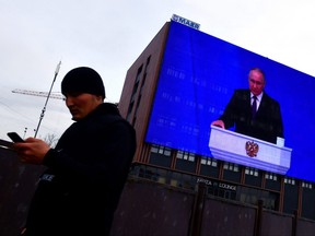 A man looks at his mobile phone as he walks past a huge screen displaying the broadcast of Russia's President Vladimir Putin's annual state of the nation address on the facade of a building in Moscow on Feb. 29, 2024.