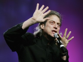 Comedian Richard Lewis gestures with his hands as he speaks into a microphone against a purple backdrop.