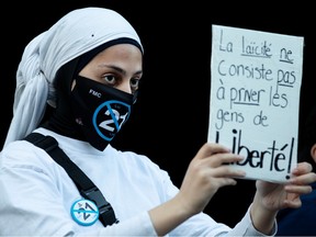 A woman in a hijab and a face mask that has "21" crossed out holds a sign that says "la laicite ne consiste pas a prener les gens de liberte"