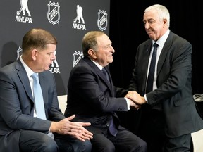 Gary Bettman sits shaking hands with a standing Luc Tardif during a news conference